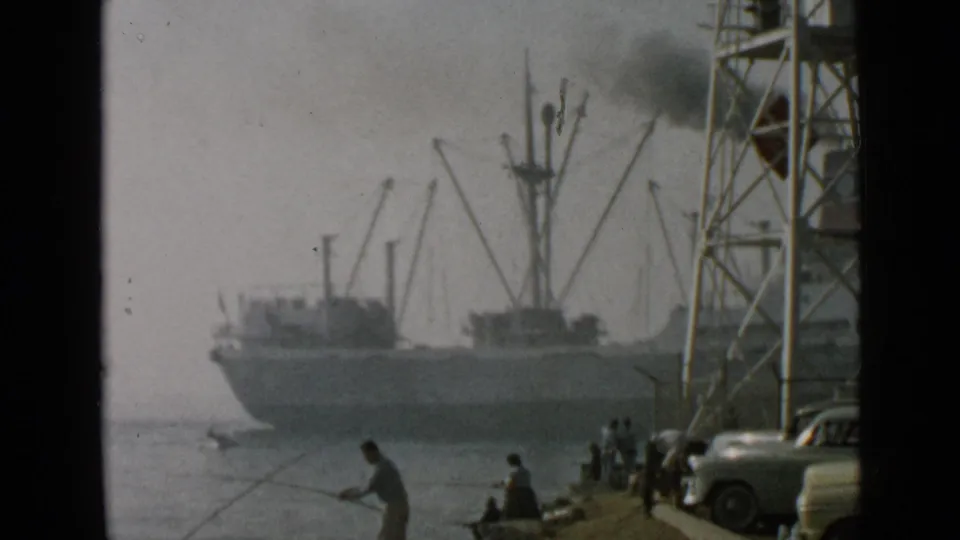 1949 San Diego harbor frame with a foreground fisherman, a smoking cargo ship, cranes, parked cars, and shoreline spectators.