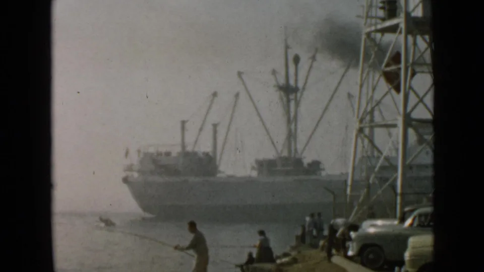 1949 San Diego harbor view with anglers on a concrete waterfront, parked cars, a metal tower, and a large ship moving through haze.