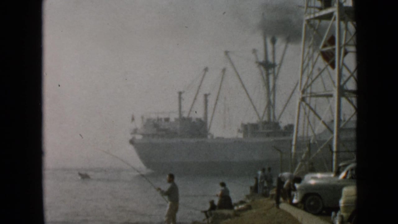 San Diego waterfront scene with a man casting, other people nearby, a metal tower, parked cars, and a large ship in the hazy harbor.