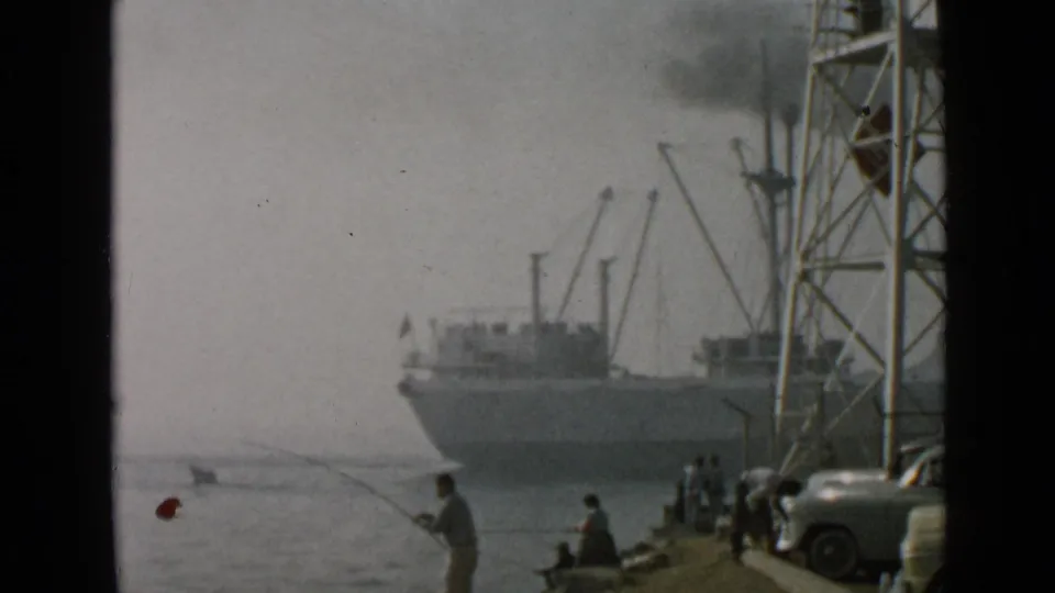 Hazy 1949 San Diego harbor footage showing people fishing from a concrete edge, with a ship, cranes, parked cars, and tower structure nearby.