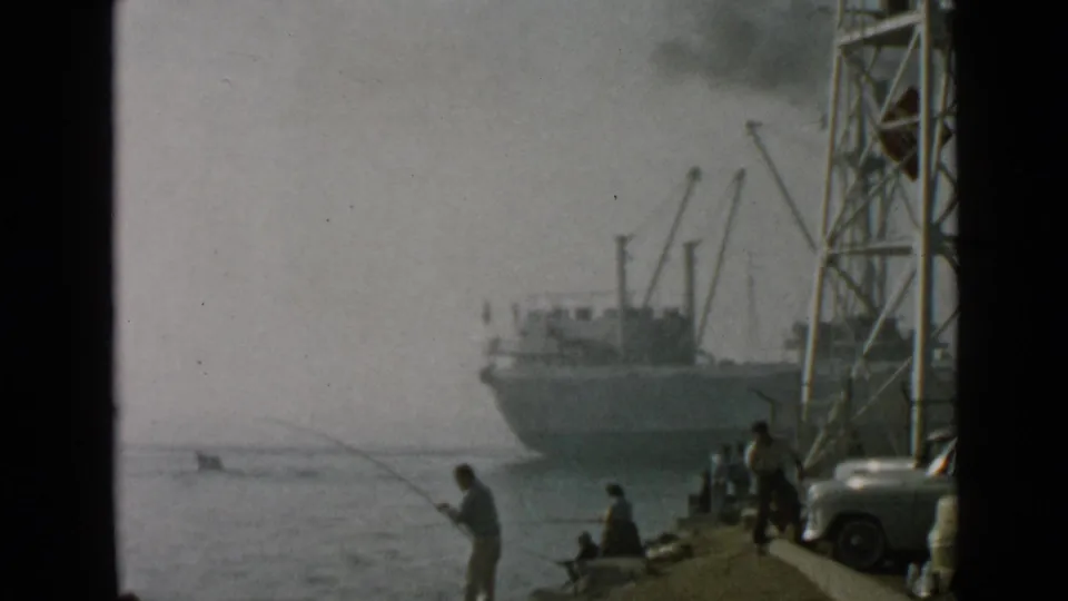 Final frame of the 1949 San Diego harbor view, with a silhouetted angler, people along the edge, parked cars, tower structure, and ship offshore.