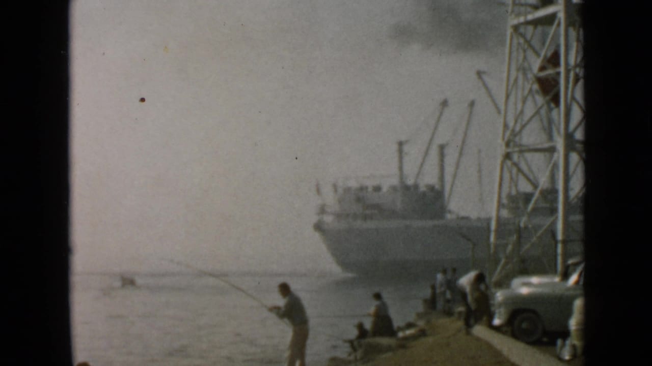 1949 San Diego home-movie frame showing a man fishing beside calm water with people seated near a docked ship and tower.