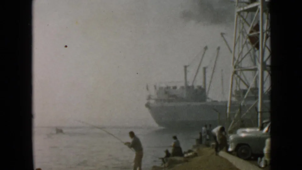 1949 San Diego home-movie frame showing a man fishing beside calm water with people seated near a docked ship and tower.
