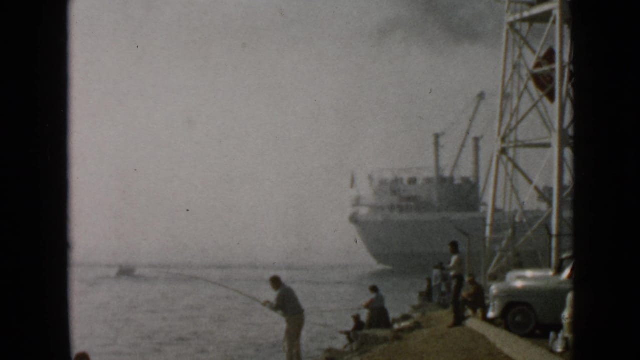 1949 San Diego waterfront frame with an angler in the foreground, bystanders on the edge, a parked car, and a docked ship.