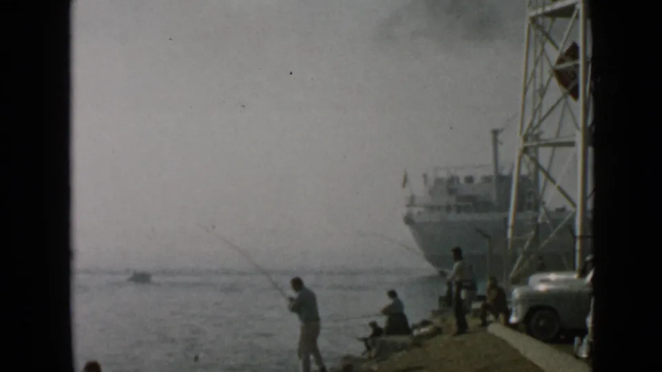1949 San Diego home-movie frame of a shoreline fishing scene with seated people, harbor structure, parked car, and ship.