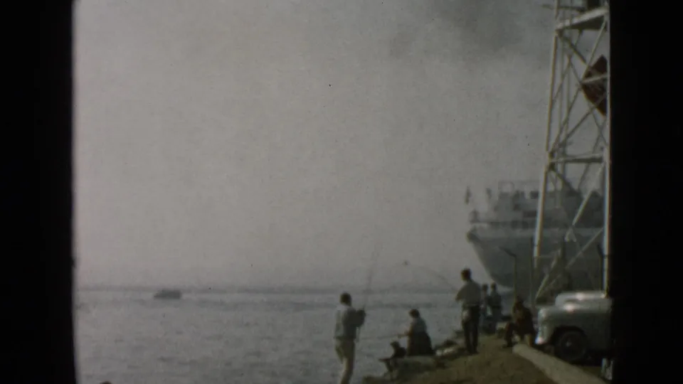 1949 San Diego harbor frame showing people on the shoreline, a fishing line over the water, a metal tower, and a large ship.