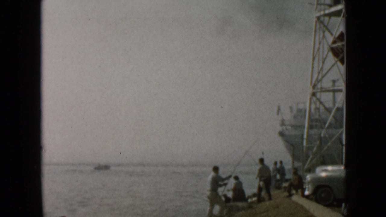 1949 San Diego home-movie frame showing anglers on a pier, a docked ship, calm water, and a rounded car at the edge.