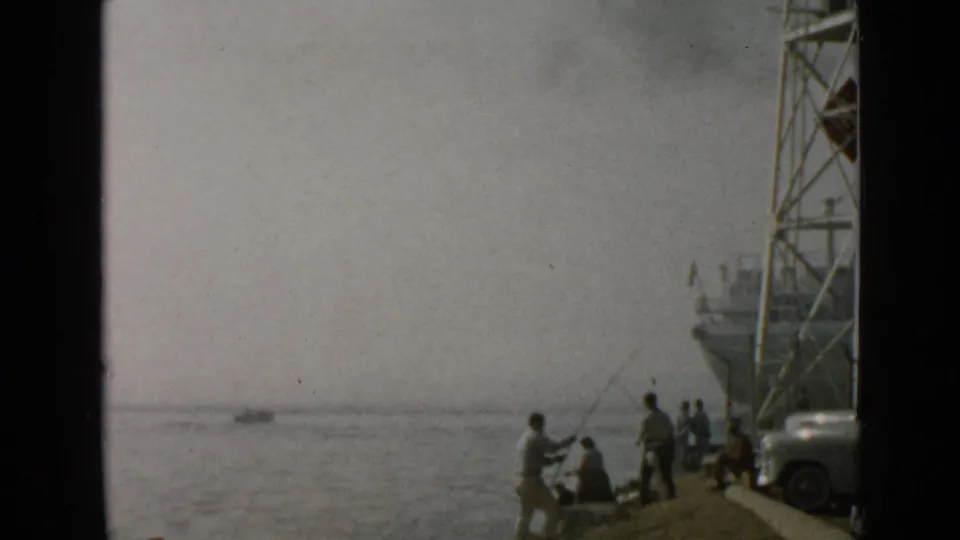 1949 San Diego home-movie frame showing anglers on a pier, a docked ship, calm water, and a rounded car at the edge.