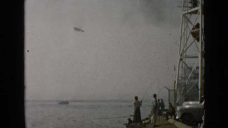 1949 San Diego pier scene with people fishing, a small boat offshore, metal tower structure, and parked automobile.