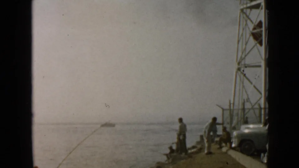 San Diego 1949 home-movie frame with anglers on a pier, an arcing fishing line, water, tower structure, and parked car.