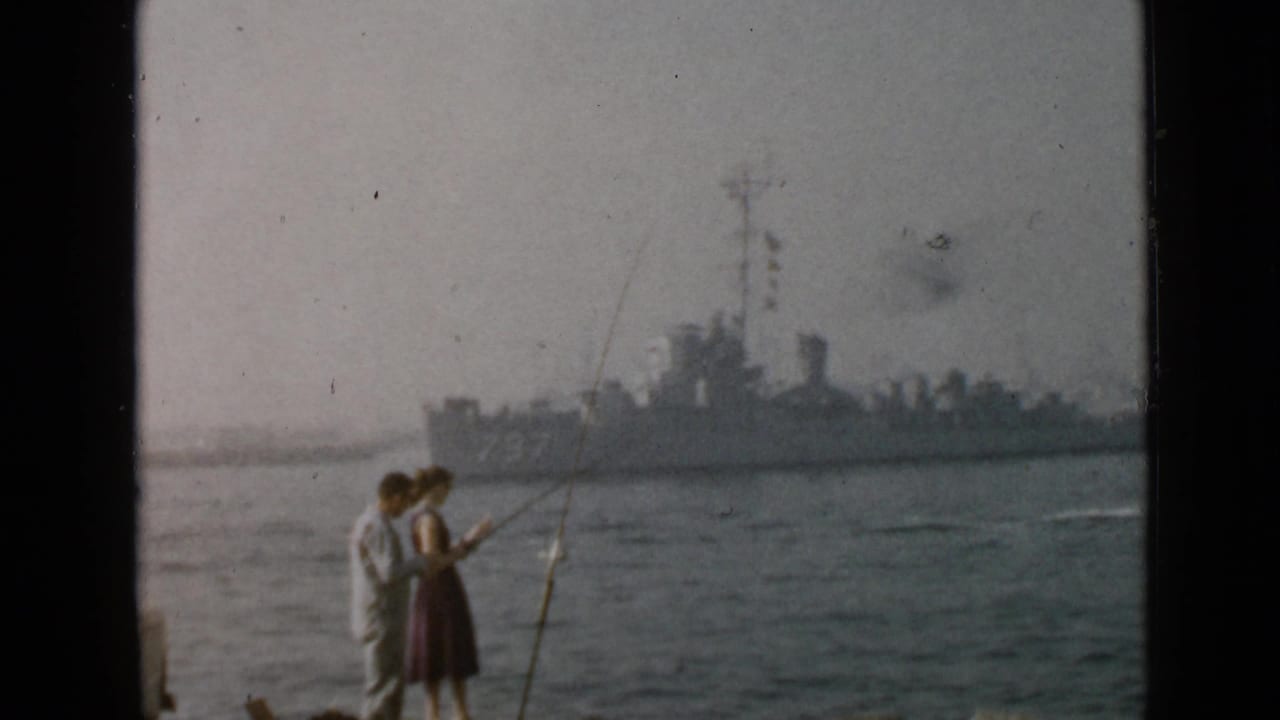 1949 San Diego home-movie frame showing a couple with fishing rods on shore as a gray warship approaches in the background.