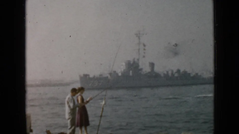1949 San Diego home-movie frame showing a couple with fishing rods on shore as a gray warship approaches in the background.