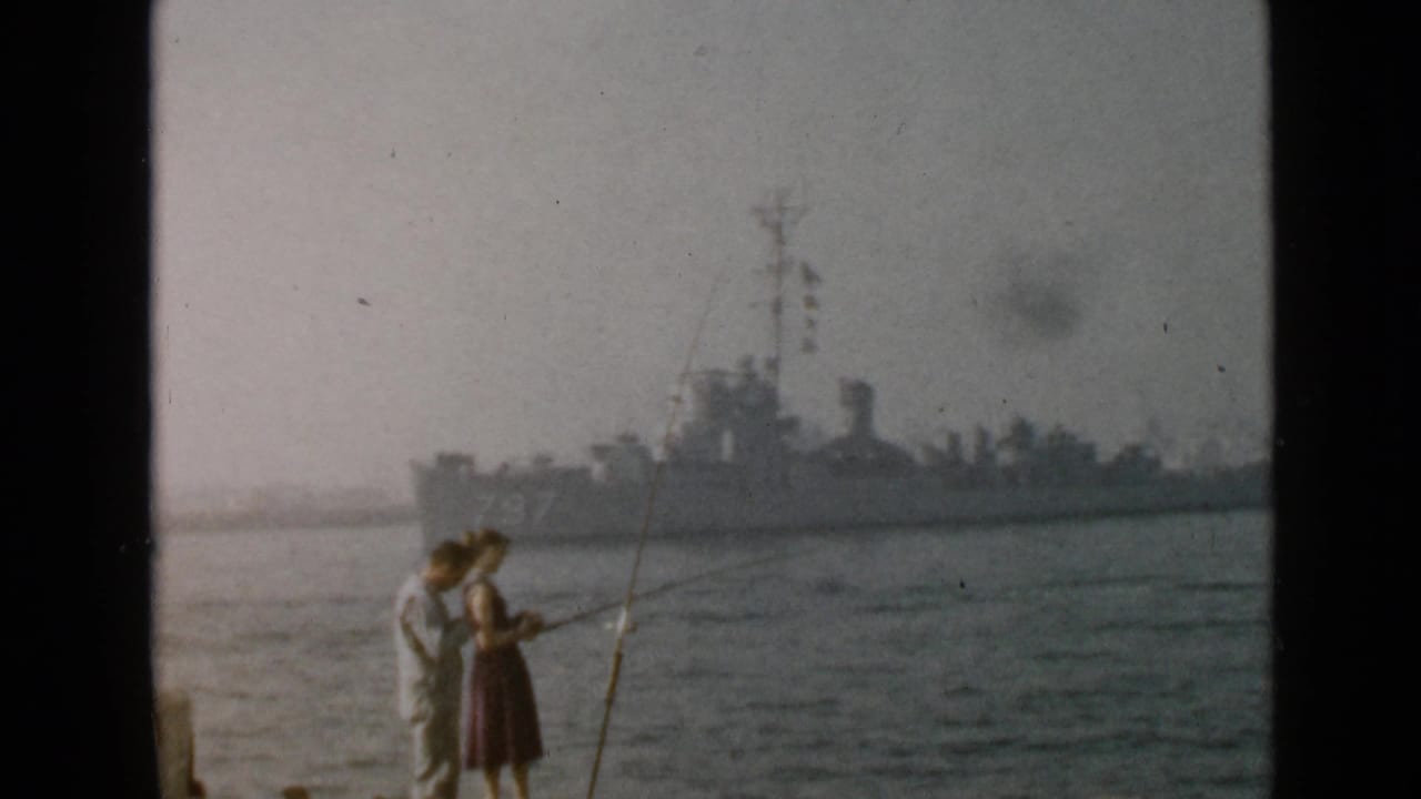 1949 San Diego frame with a man and woman fishing near the water while a gray naval ship marked 797 is visible behind them.