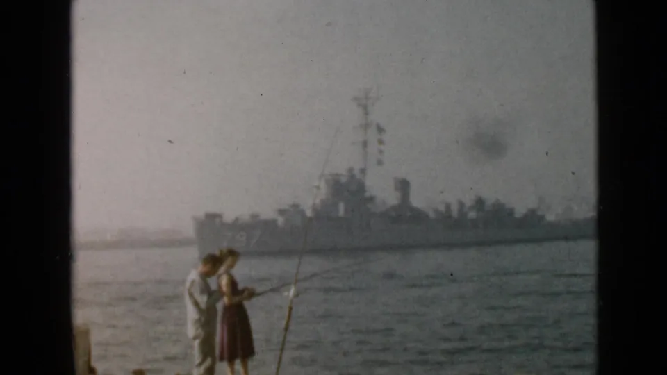 1949 San Diego frame with a man and woman fishing near the water while a gray naval ship marked 797 is visible behind them.