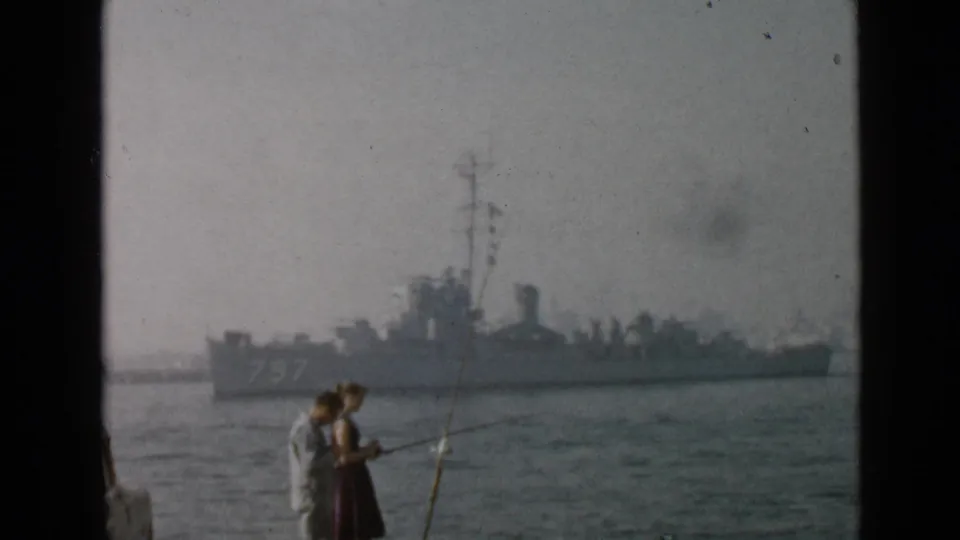 Late-1940s San Diego harbor frame with a couple fishing from shore and a gray naval warship marked 797 in the distance.