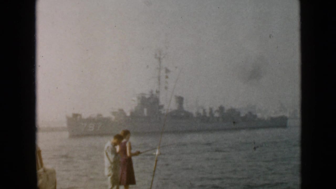In 1949 San Diego footage, a man and woman stand with fishing rods near the water as a naval ship passes in the distance.