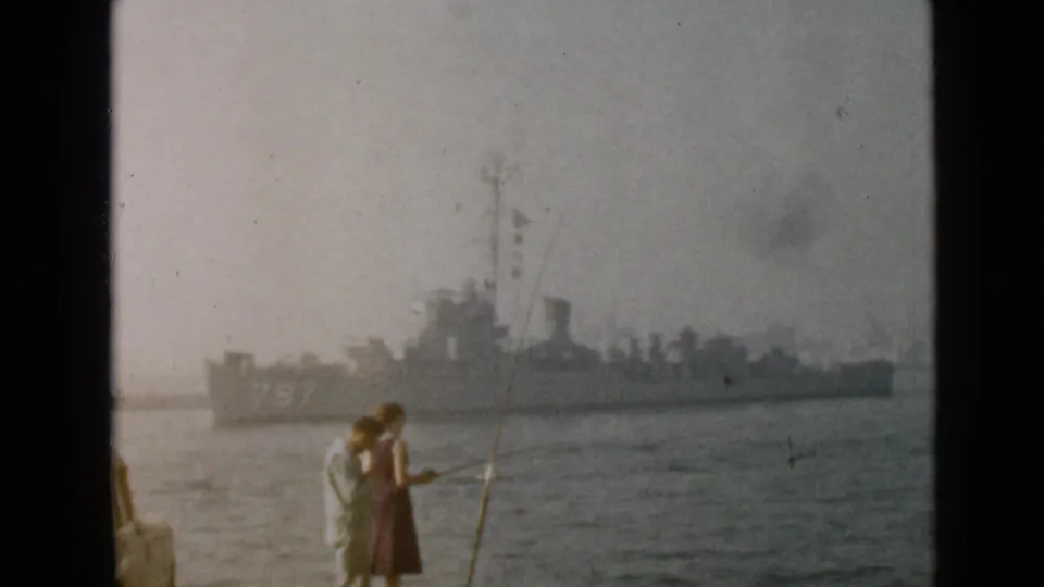 In 1949 San Diego footage, a man and woman stand with fishing rods near the water as a naval ship passes in the distance.