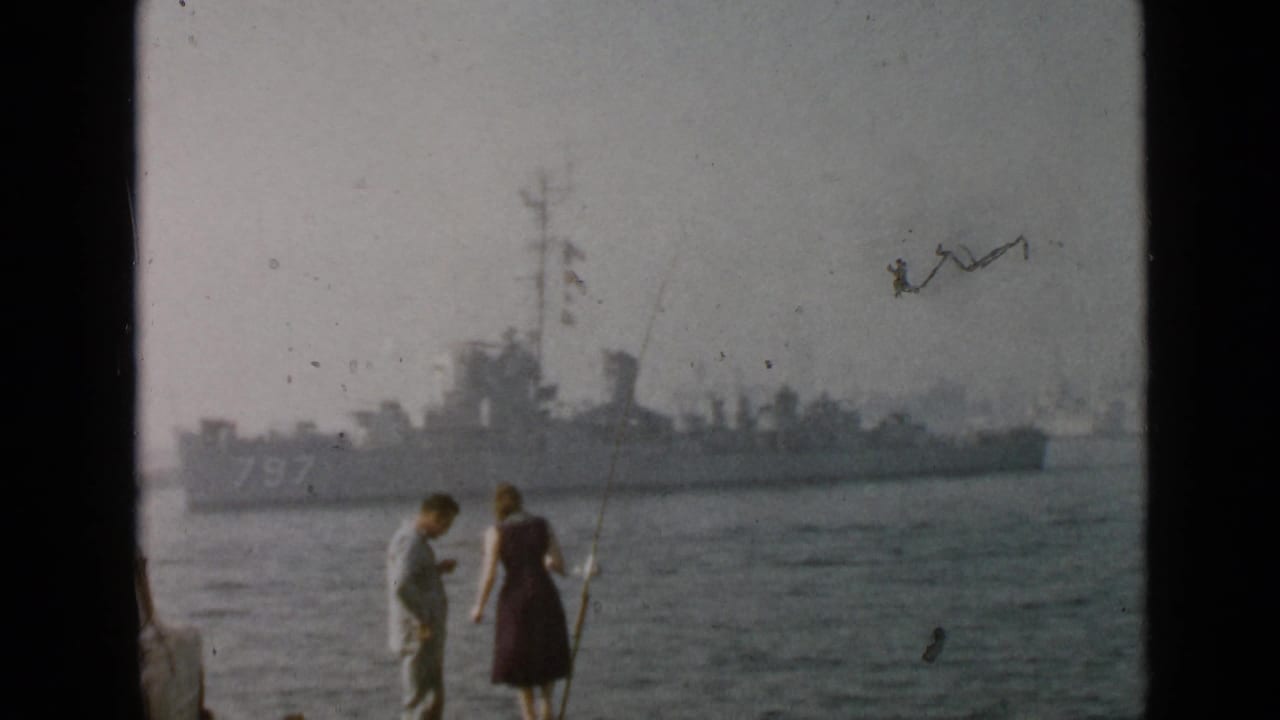 A man and woman fish along the San Diego waterfront while a distant naval vessel marked 797 is visible across the water.