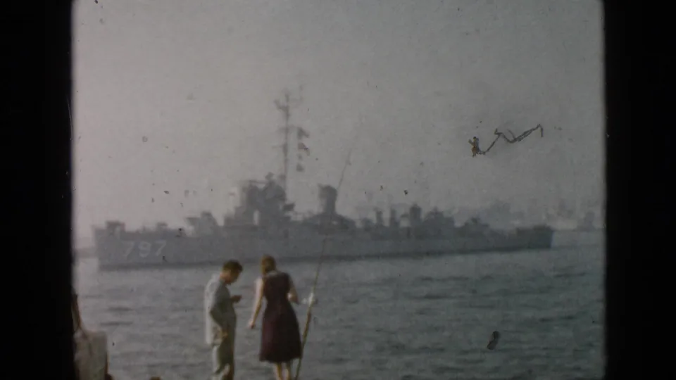 A man and woman fish along the San Diego waterfront while a distant naval vessel marked 797 is visible across the water.