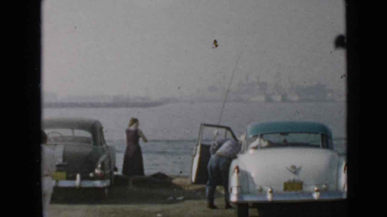 At the San Diego harbor shore, two adults are near a white classic car and a dark sedan with water and a distant skyline behind them.