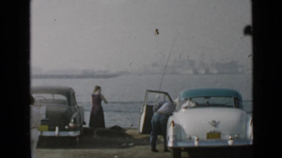 At the San Diego harbor shore, two adults are near a white classic car and a dark sedan with water and a distant skyline behind them.