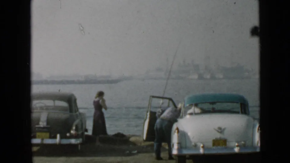 A woman in a dark dress stands at the San Diego harbor shore as a man bends into a white classic car beside another parked sedan.