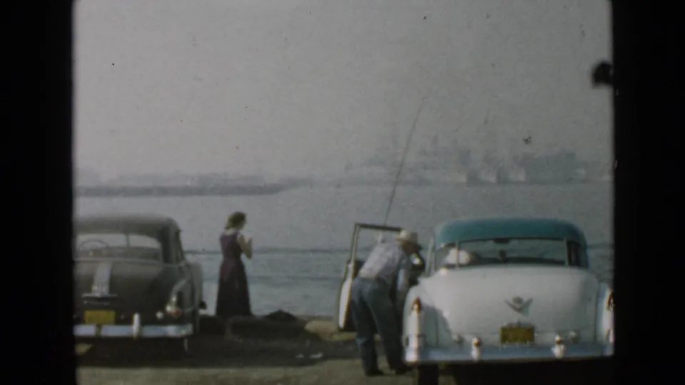 Near the San Diego harbor, a man bends into the open door of a white classic car while a woman stands by the water in the background.