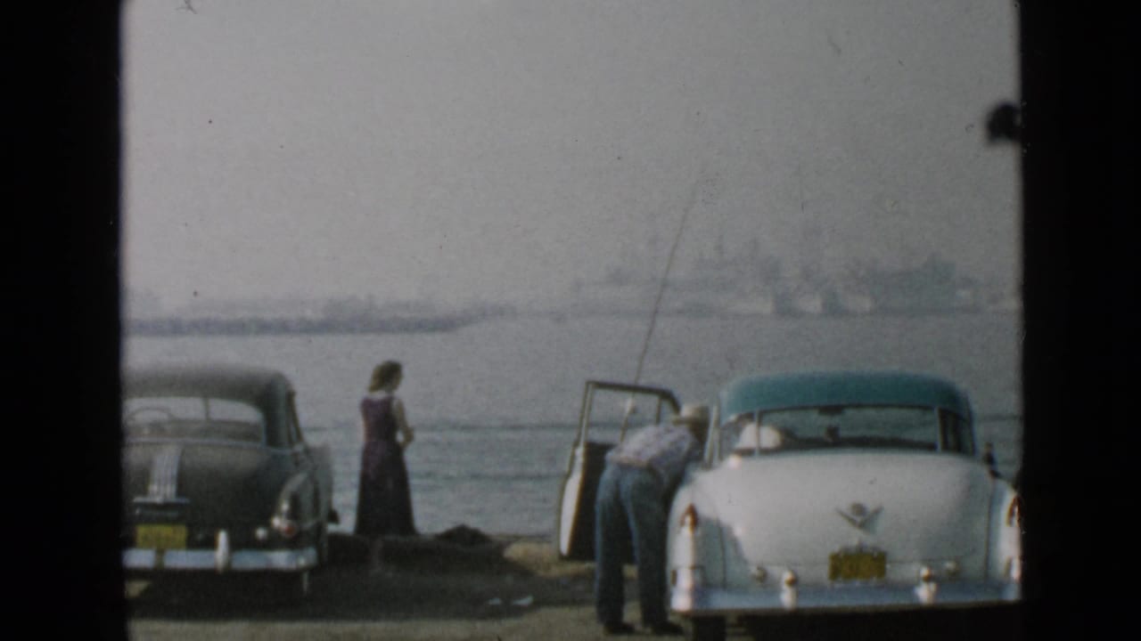 1949 San Diego home-movie frame showing two adults near parked cars at the waterfront, with fishing rods and a distant shoreline visible.