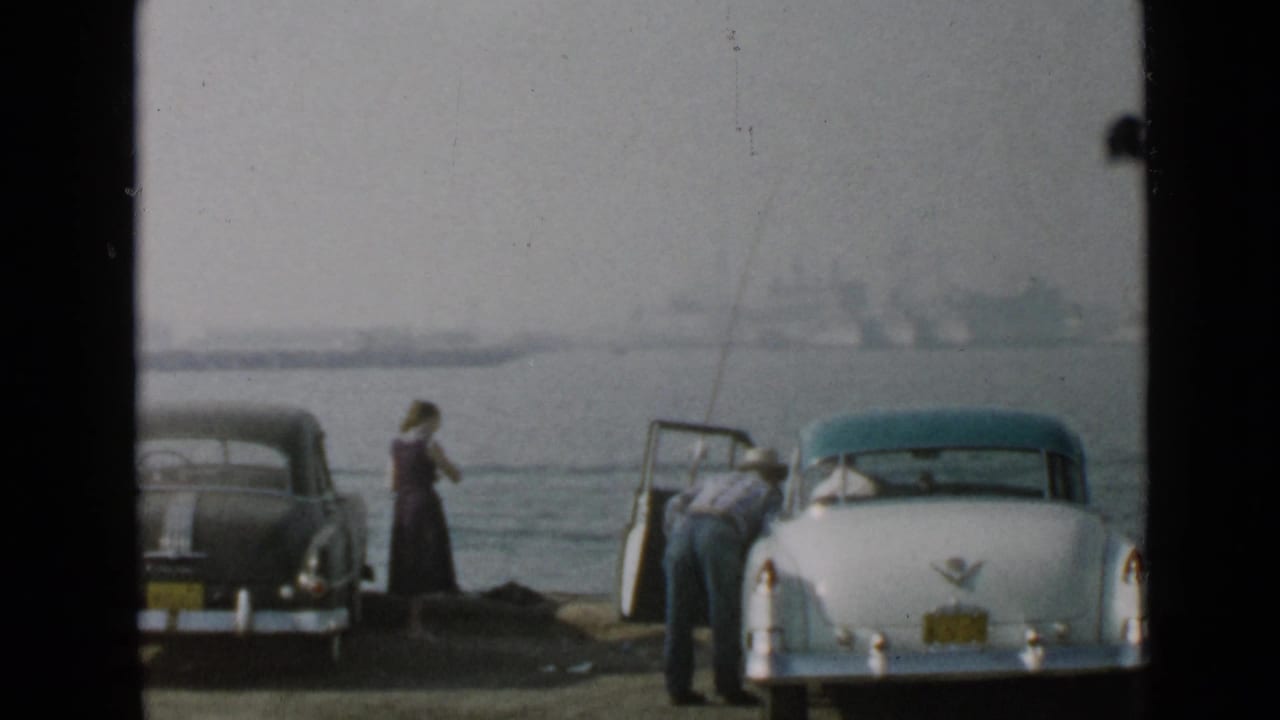1949 San Diego waterfront frame with a woman standing near the water and a man at an open white car door beside fishing equipment.