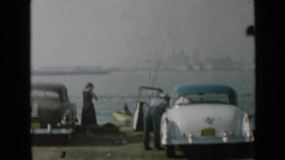 1949 San Diego home-movie frame showing two adults by parked cars at the shoreline, with fishing gear and a small boat near the water.