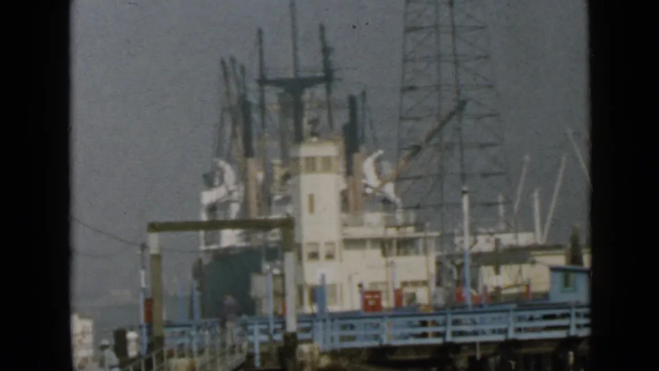 A docked cargo ship in 1949 San Diego is seen from the pier, with blue railings, a distant walking figure, rigging, and harbor equipment.