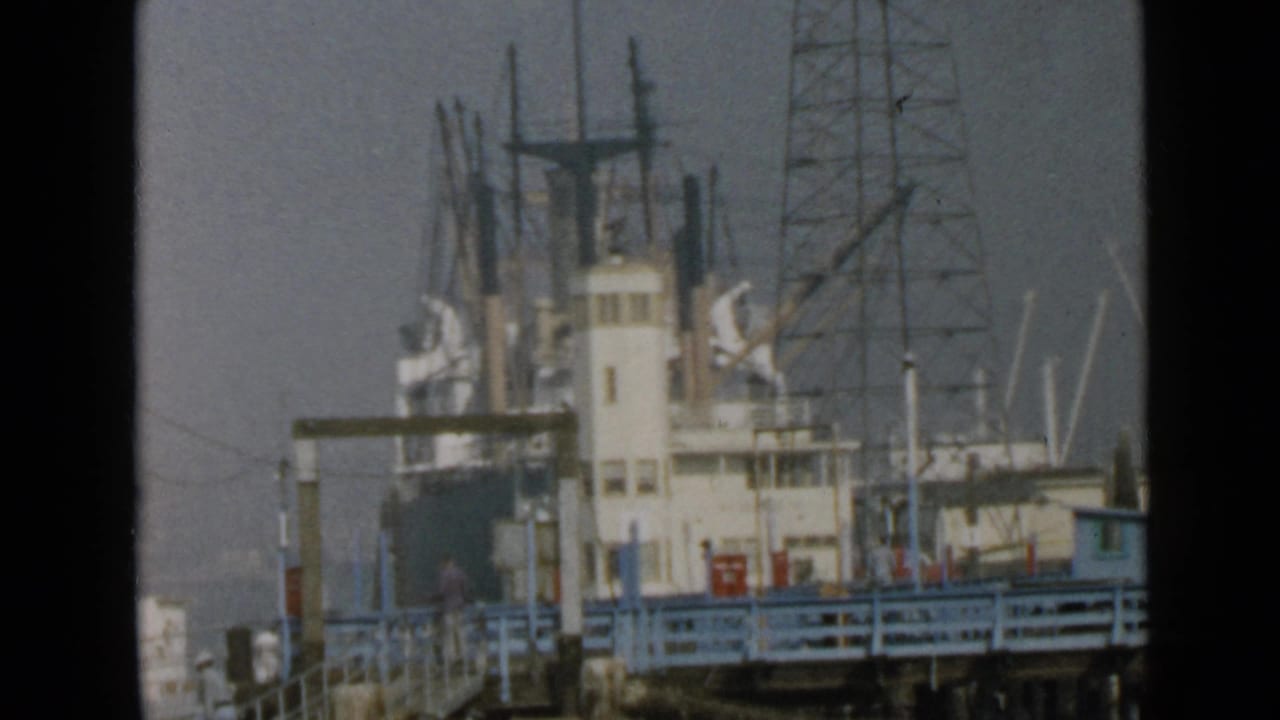 1949 San Diego waterfront view of a docked cargo ship behind blue pier railings, filmed in soft 8mm color.