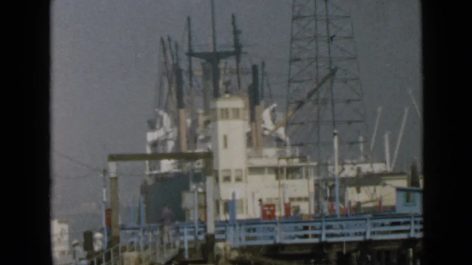 1949 San Diego waterfront view of a docked cargo ship behind blue pier railings, filmed in soft 8mm color.