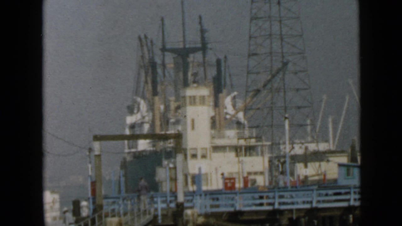 Similar harbor view of docked ship, mast rigging, tower-like structure, and pier railing.