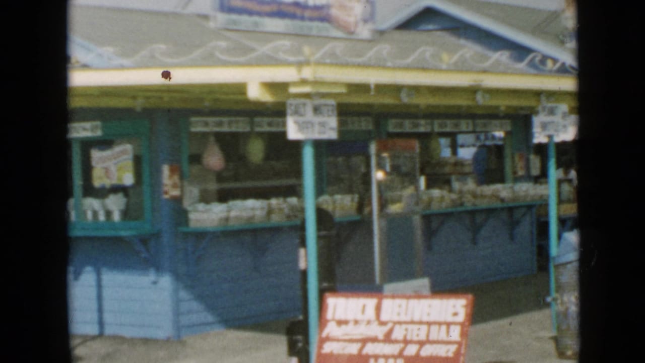 1949 San Diego 8mm frame showing a blue food stand with yellow trim, counter signage, and a red delivery sign near the foreground.