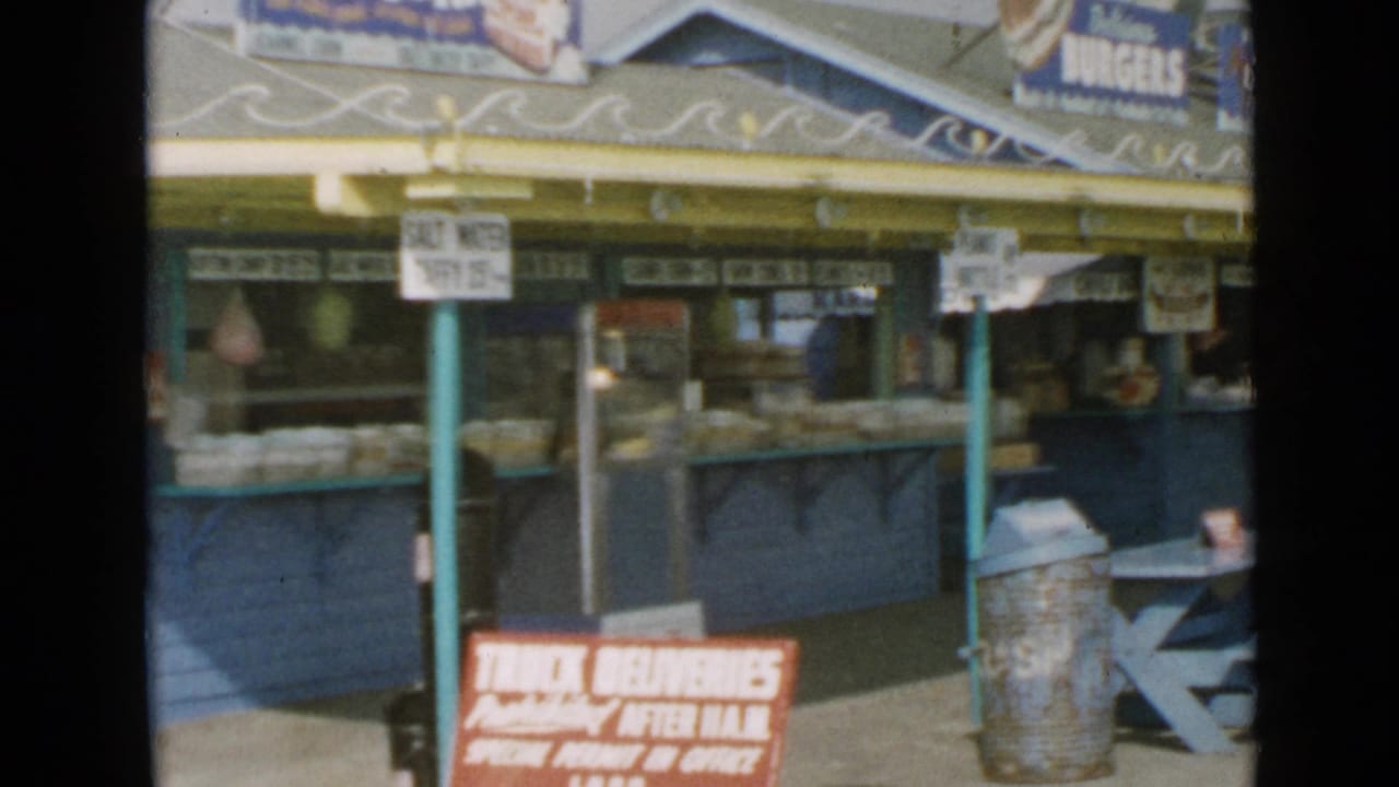 Food stand counter with burger signage, trash can, and picnic table at right.