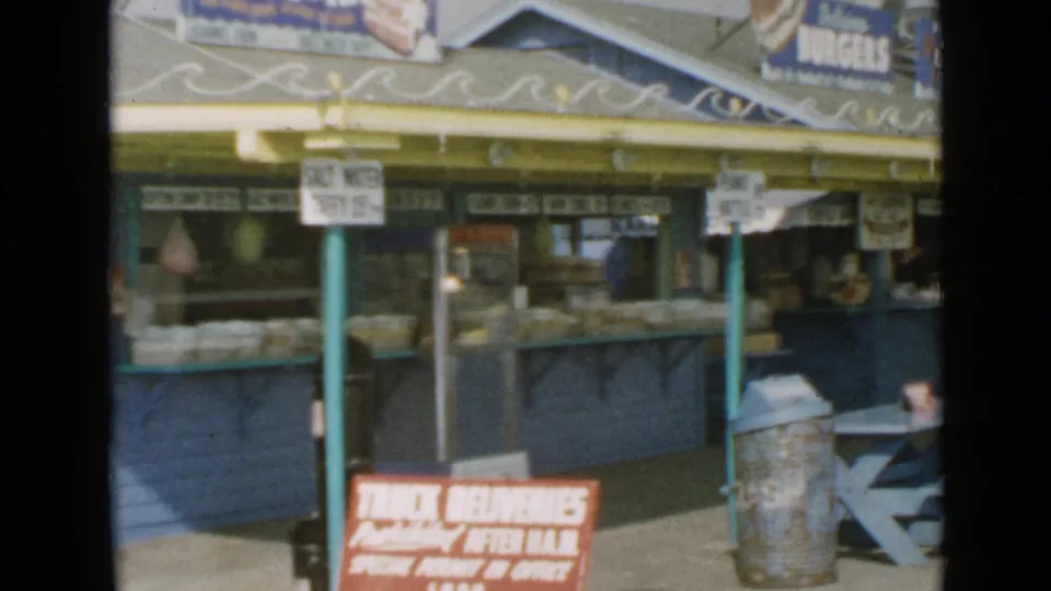 1949 San Diego home-movie frame with a snack counter, burger sign, metal trash can, and blue picnic table seating.