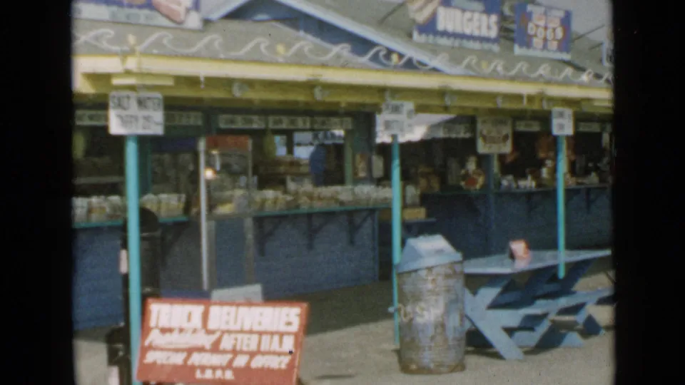 1949 San Diego 8mm view along the outdoor food stand, showing counter displays, signage, trash can, and picnic seating.