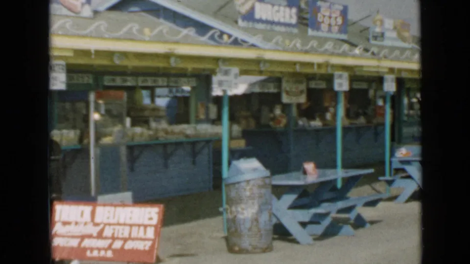 End of the 1949 San Diego food-stand pan, with picnic tables, a metal trash can, counter windows, and overhead snack signs.
