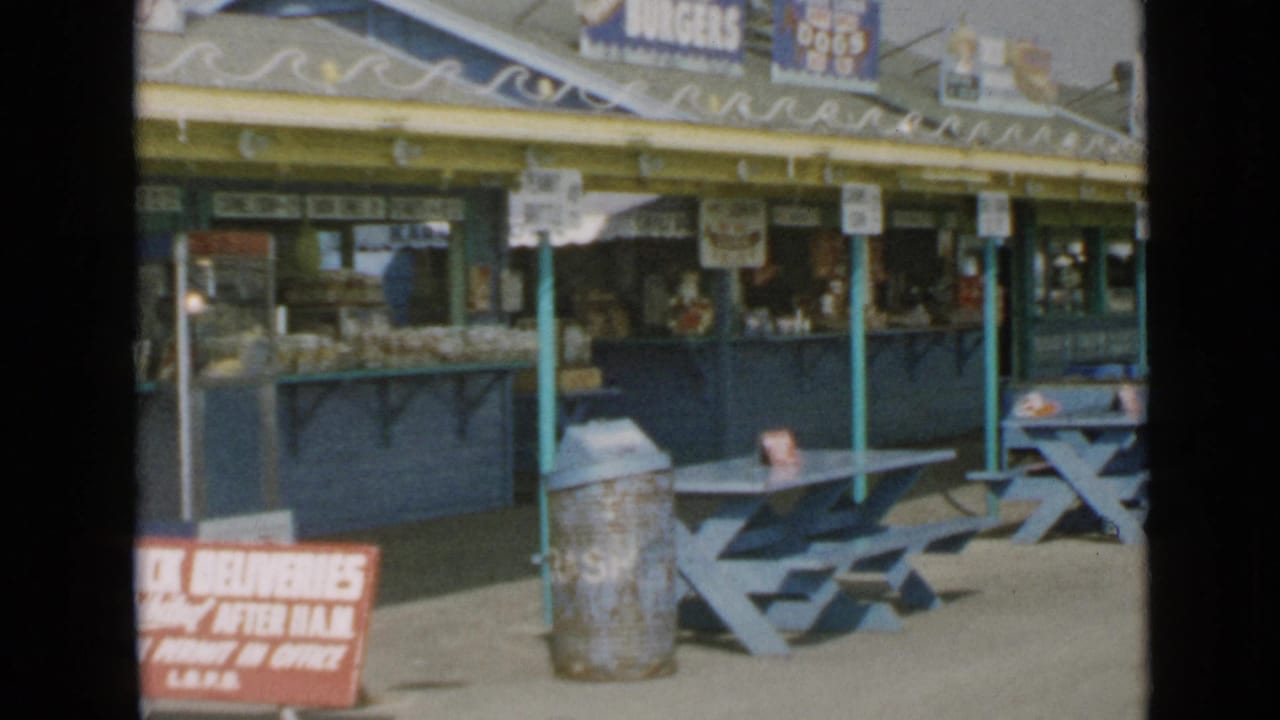 1949 San Diego boardwalk food stand with blue picnic tables, a metal trash can, yellow awning, and hanging food signs.
