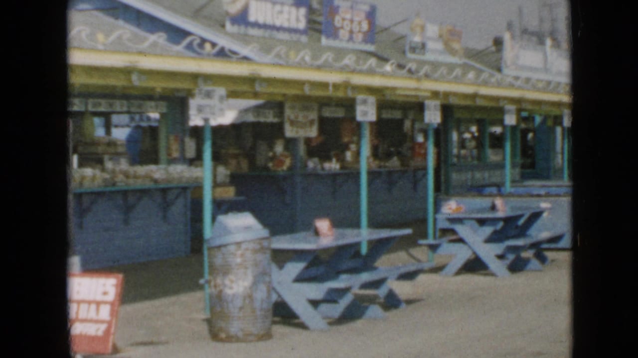 Mid-century San Diego boardwalk concession area with blue tables, metal trash can, food counter, awning, and rooftop signs.