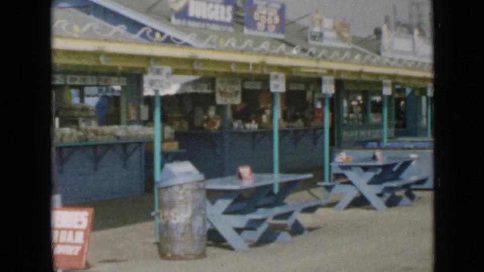 Mid-century San Diego boardwalk concession area with blue tables, metal trash can, food counter, awning, and rooftop signs.