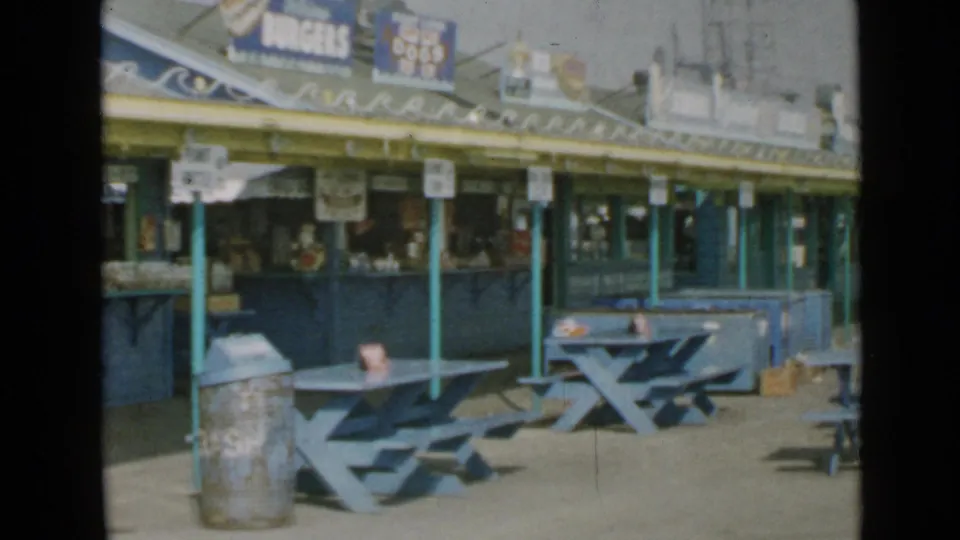 1949 San Diego boardwalk scene showing blue picnic tables, concession windows, painted posts, and food signs under a yellow awning.