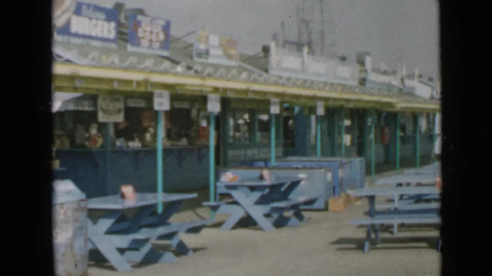 End of a 1949 San Diego boardwalk pan showing a long concession stand, blue picnic tables, railing, awning, and food signage.
