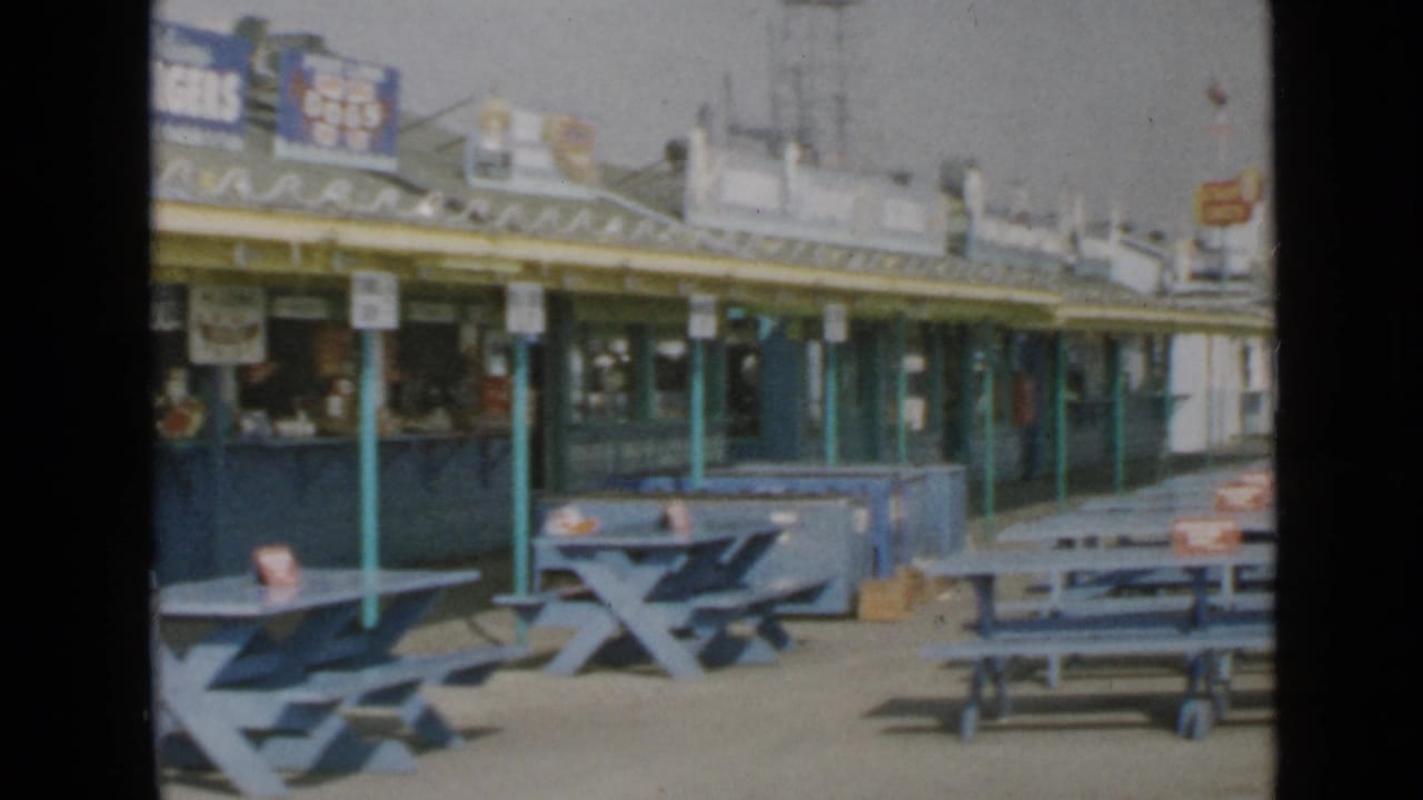 1949 San Diego pier scene with blue outdoor seating, green support poles, and food-stand counters under a sunny sky.