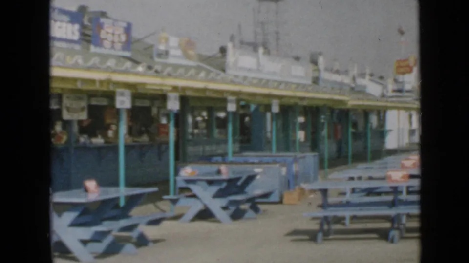 1949 San Diego pier scene with blue outdoor seating, green support poles, and food-stand counters under a sunny sky.