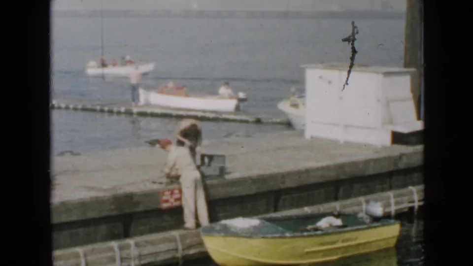 1949 San Diego waterfront frame showing a person on a pier, a yellow boat edge, and small boats near a dock.