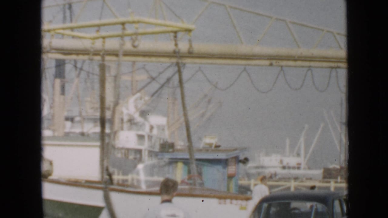 Small boat near a dock crane with fishing vessels and cars in the harbor background.