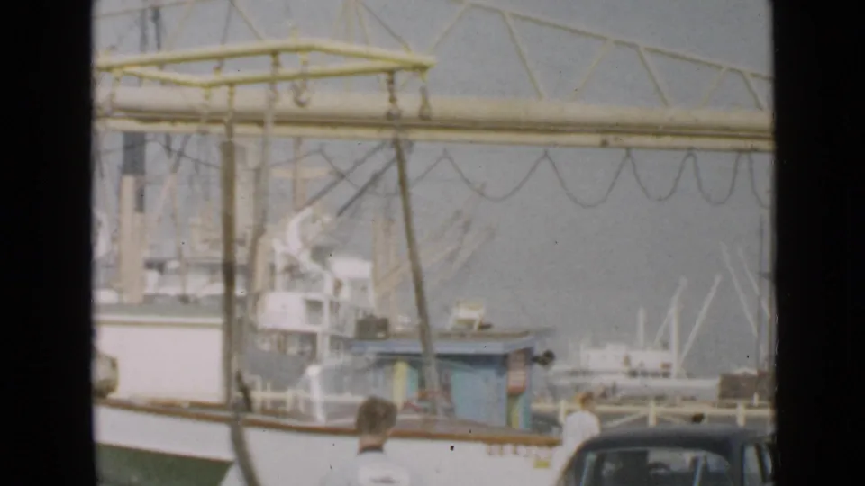 1949 San Diego harbor view with a small boat by a yellow gantry crane, larger fishing boats, dock structures, and older cars visible.