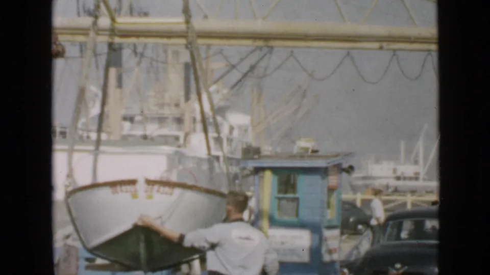 A man in a light shirt guides a small boat hanging from a yellow gantry crane at the San Diego harbor dock in 1949.